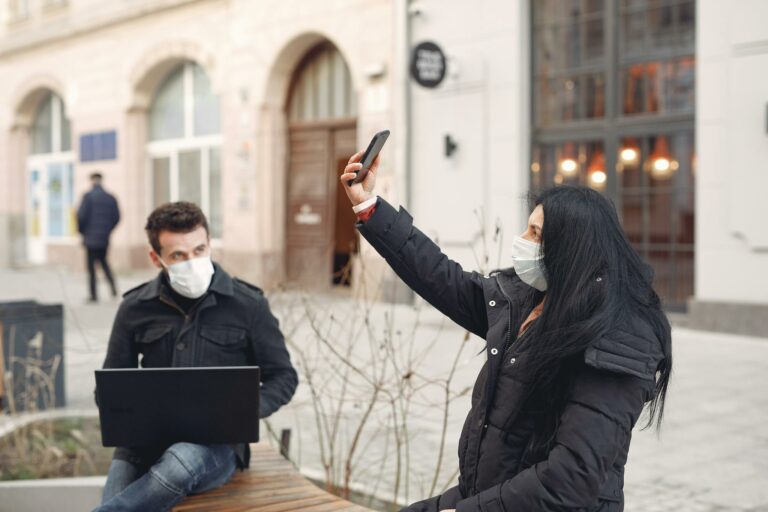Young couple wearing medical masks using laptop and smartphone on city street